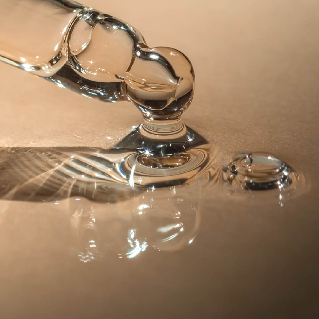 Nahaufnahme einer Glaspipette, die einen Flüssigkeitstropfen auf eine glatte Oberfläche abgibt. Sowohl die klare Flüssigkeit als auch die Pipette reflektieren das Licht und erzeugen komplizierte Muster und Reflexionen. Der Hintergrund ist sanft beleuchtet, wodurch die Transparenz der Pipette und der Flüssigkeit hervorgehoben wird. Nahaufnahme einer Glaspipette, die einen Flüssigkeitstropfen auf eine glatte Oberfläche abgibt. Sowohl die klare Flüssigkeit als auch die Pipette reflektieren das Licht und erzeugen komplizierte Muster und Reflexionen. Der Hintergrund ist sanft beleuchtet, wodurch die Transparenz der Pipette und der Flüssigkeit hervorgehoben wird.