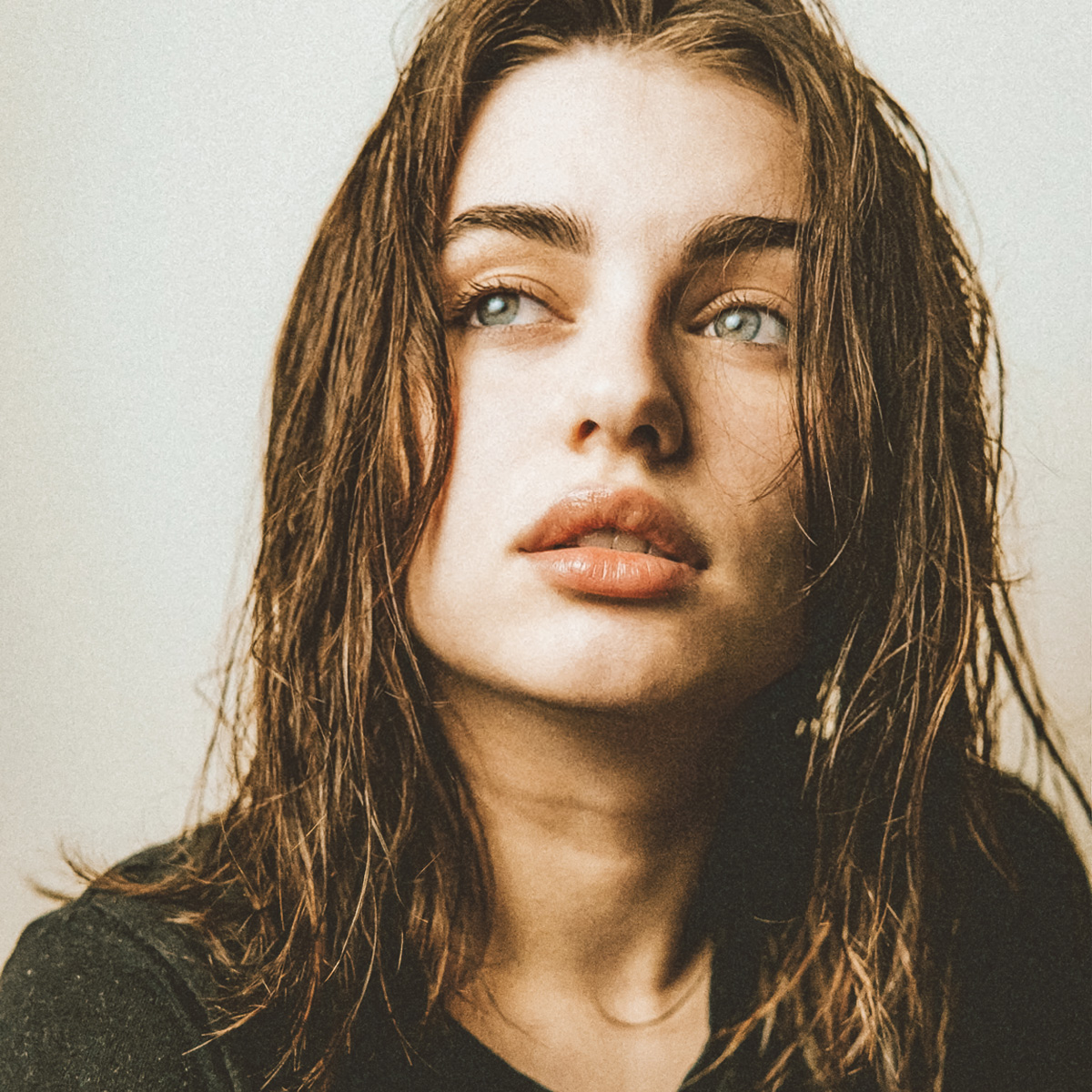 Eine Nahaufnahme einer jungen Frau mit nassem, schulterlangem Haar, die leicht nach oben und zur Seite blickt. Sie hat helle Augen, volle Lippen und trägt ein dunkles Oberteil. Der Hintergrund ist schlicht und in einem neutralen Ton gehalten. Eine Nahaufnahme einer jungen Frau mit nassem, schulterlangem Haar, die leicht nach oben und zur Seite blickt. Sie hat helle Augen, volle Lippen und trägt ein dunkles Oberteil. Der Hintergrund ist schlicht und in einem neutralen Ton gehalten.